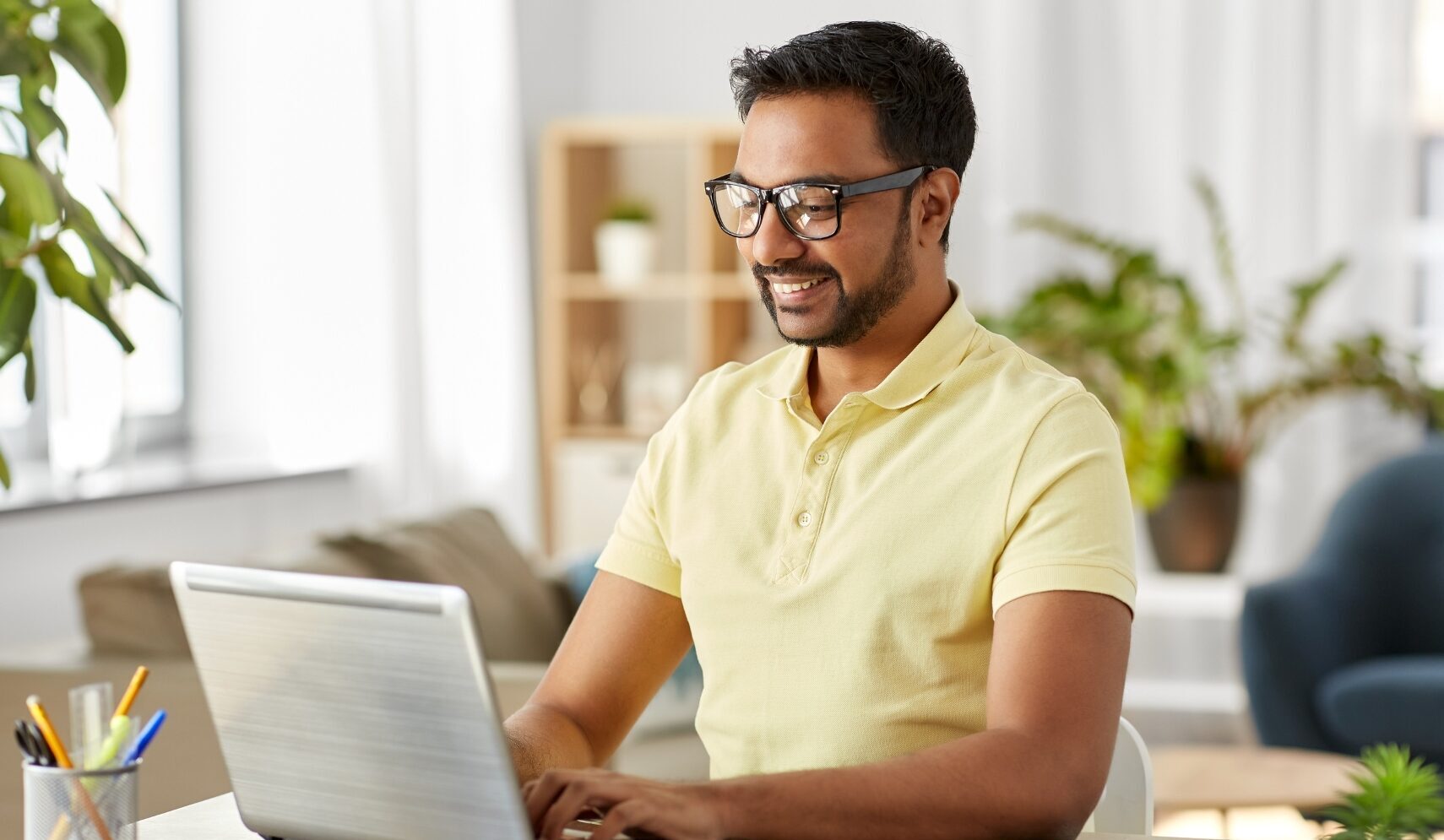 Man in yellow polo shirt smiling while sitting at a table and working on his laptop.