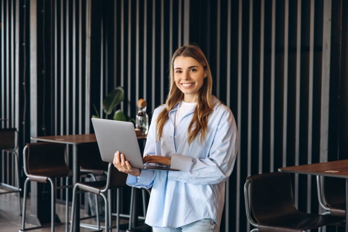 A young professional standing with a laptop in a modern office, representing new sales career opportunities and the start of a sales career journey.
