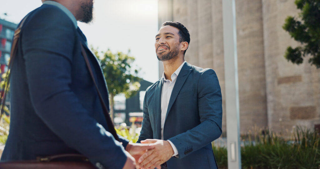 Sales professional sales trainer is need shaking hands with a sales client and a technique to get to know his prospect. They are outside and both wearing suits.