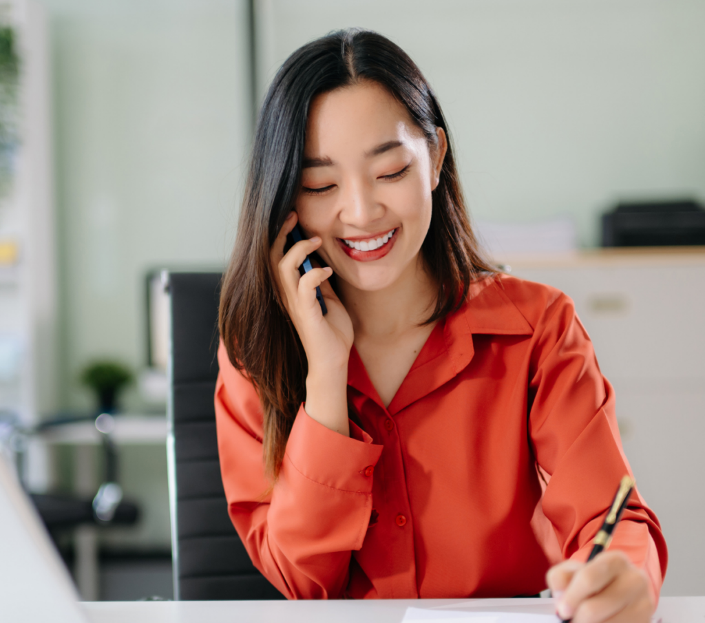 Sales professional speaking with a client by phone while taking notes at her desk.