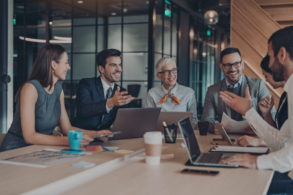 Temporary Sales Hiring - image of sales team sitting around conference table with computers open and laughing during meeting
